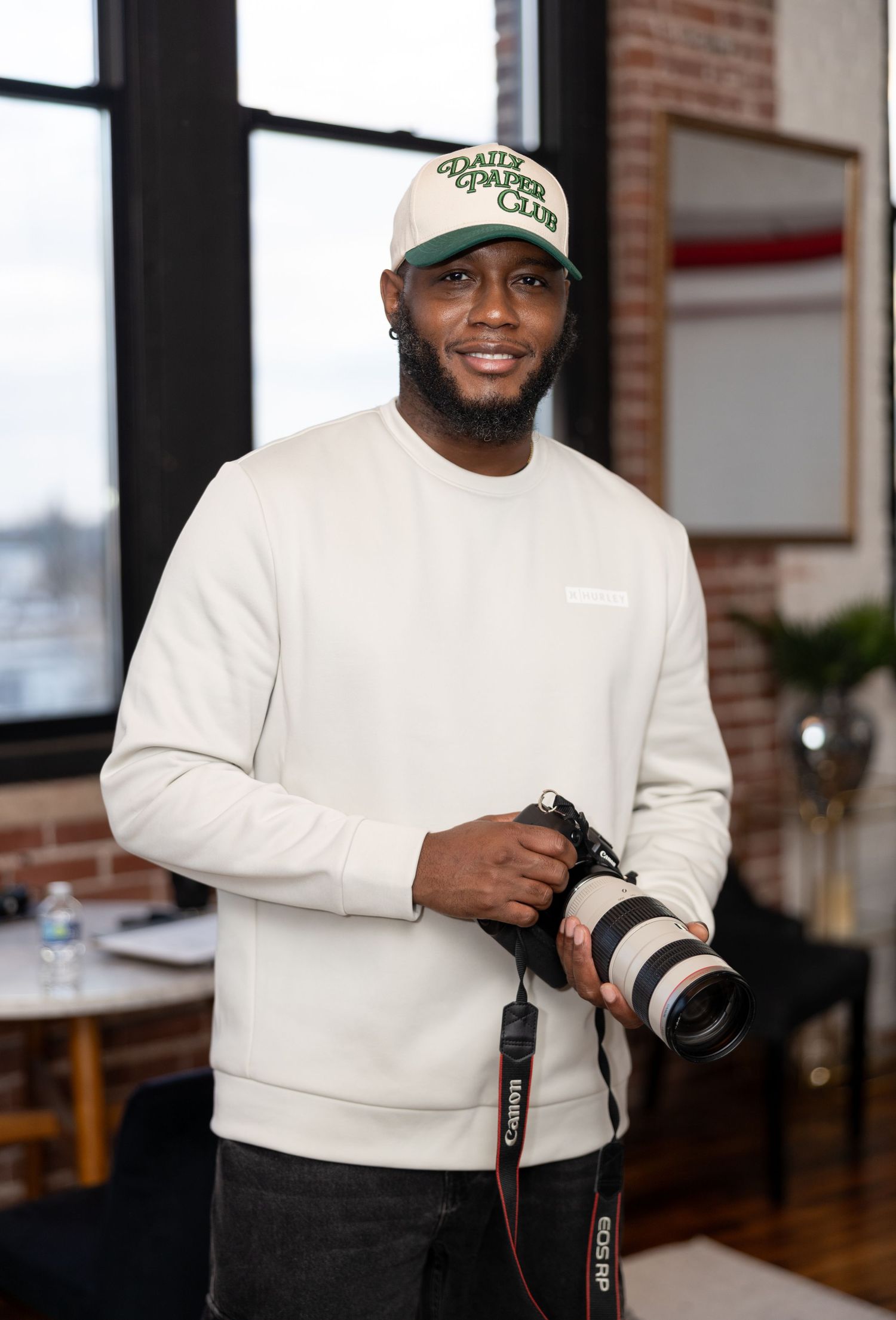 man standing indoors, wearing a white long-sleeved shirt, dark pants, and a baseball cap. He is holding a professional camera.