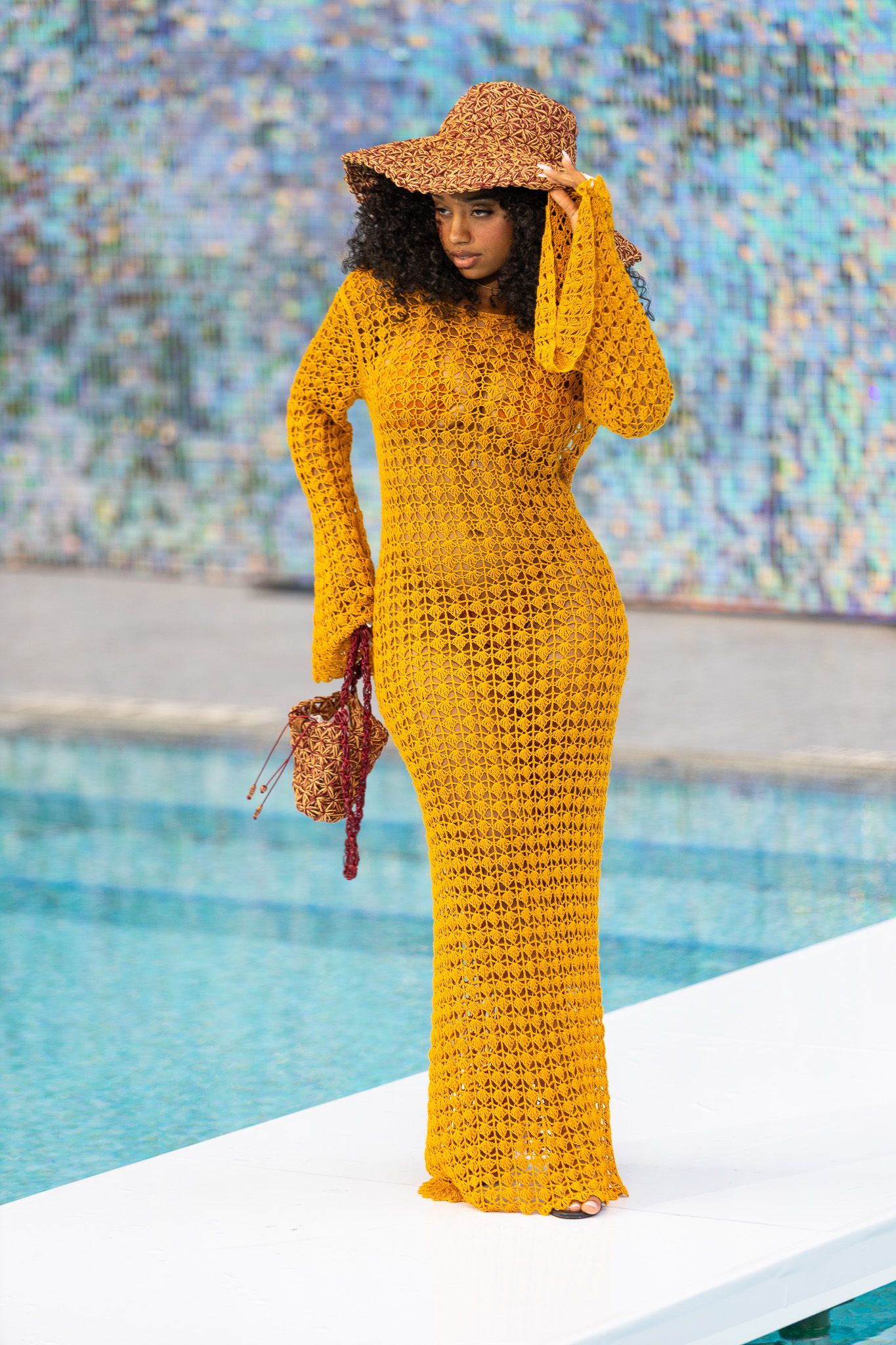 This photo features a woman standing outdoors, wearing a long, yellow, long-sleeved dress and a wide-brimmed hat.