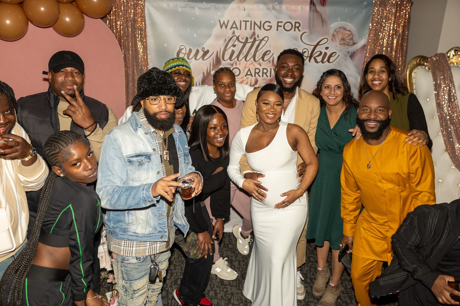 This photo features a group of people posing together indoors, likely at a celebration. A pregnant woman in a white dress stands.
