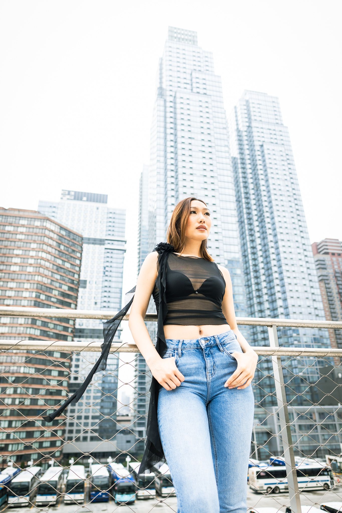 a woman standing on a bridge with a cityscape in the background. She is wearing a black top and blue jeans, and the scene is brightly lit.