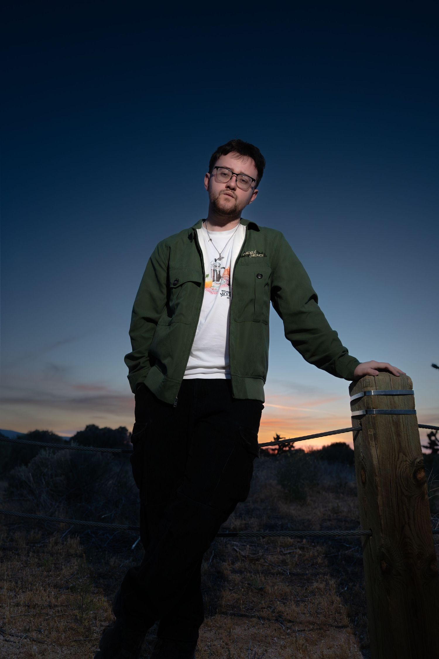 Young man in green jacket leans on wooden fence post at dusk with colorful sunset sky behind him.