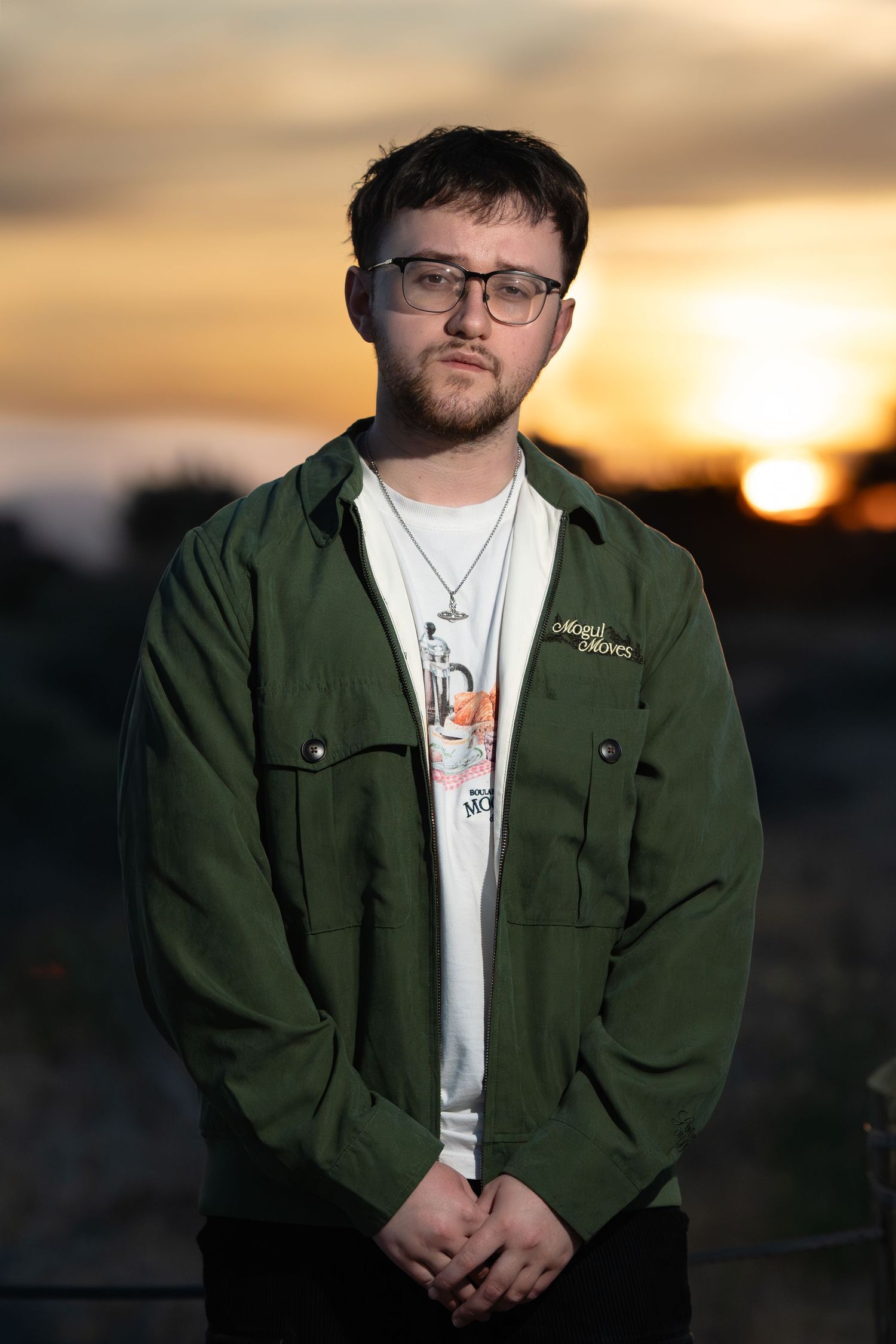 Young man in green jacket and glasses standing outdoors during golden hour sunset.