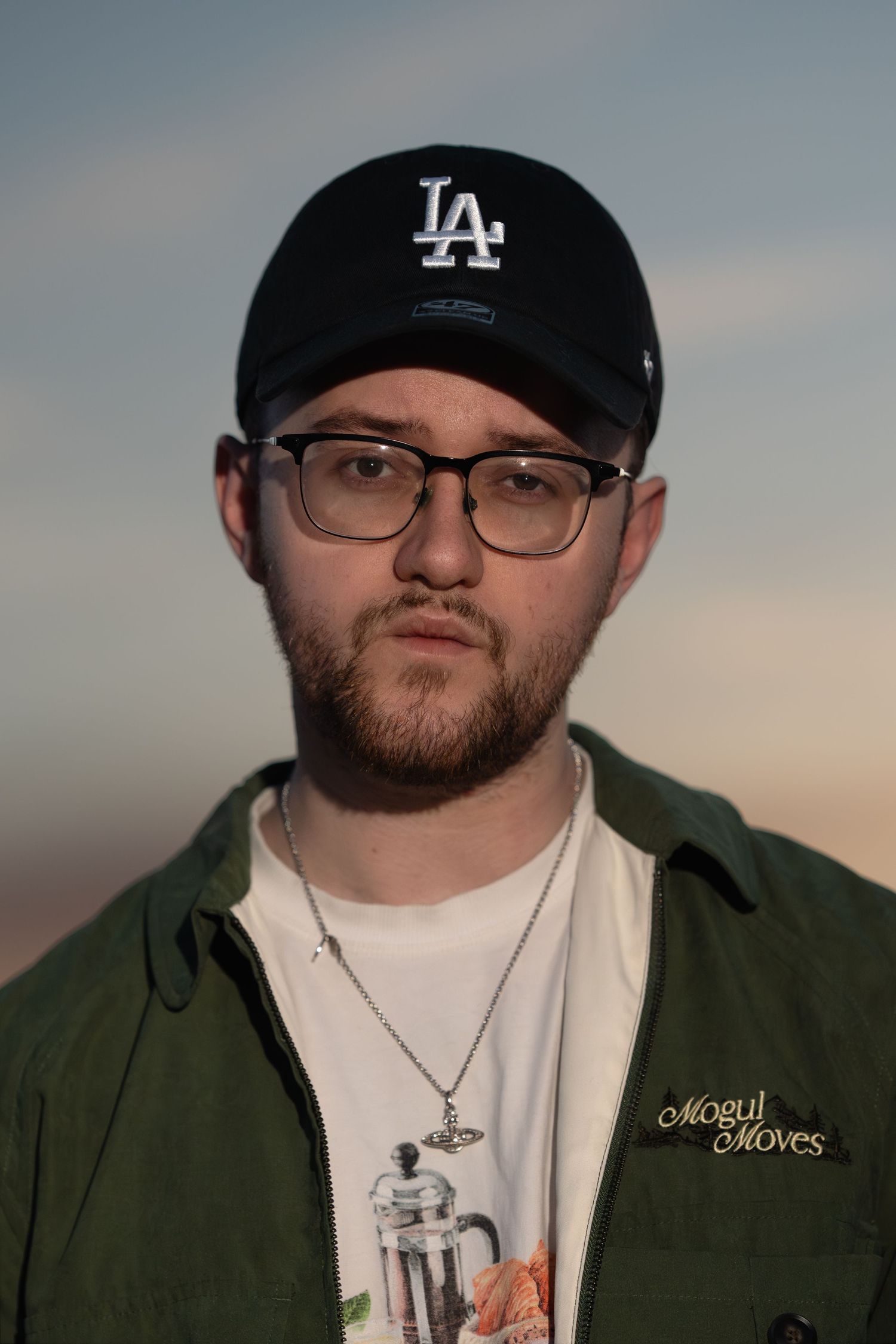 Young man with beard and glasses wearing LA Dodgers cap and green jacket outdoors at dusk.