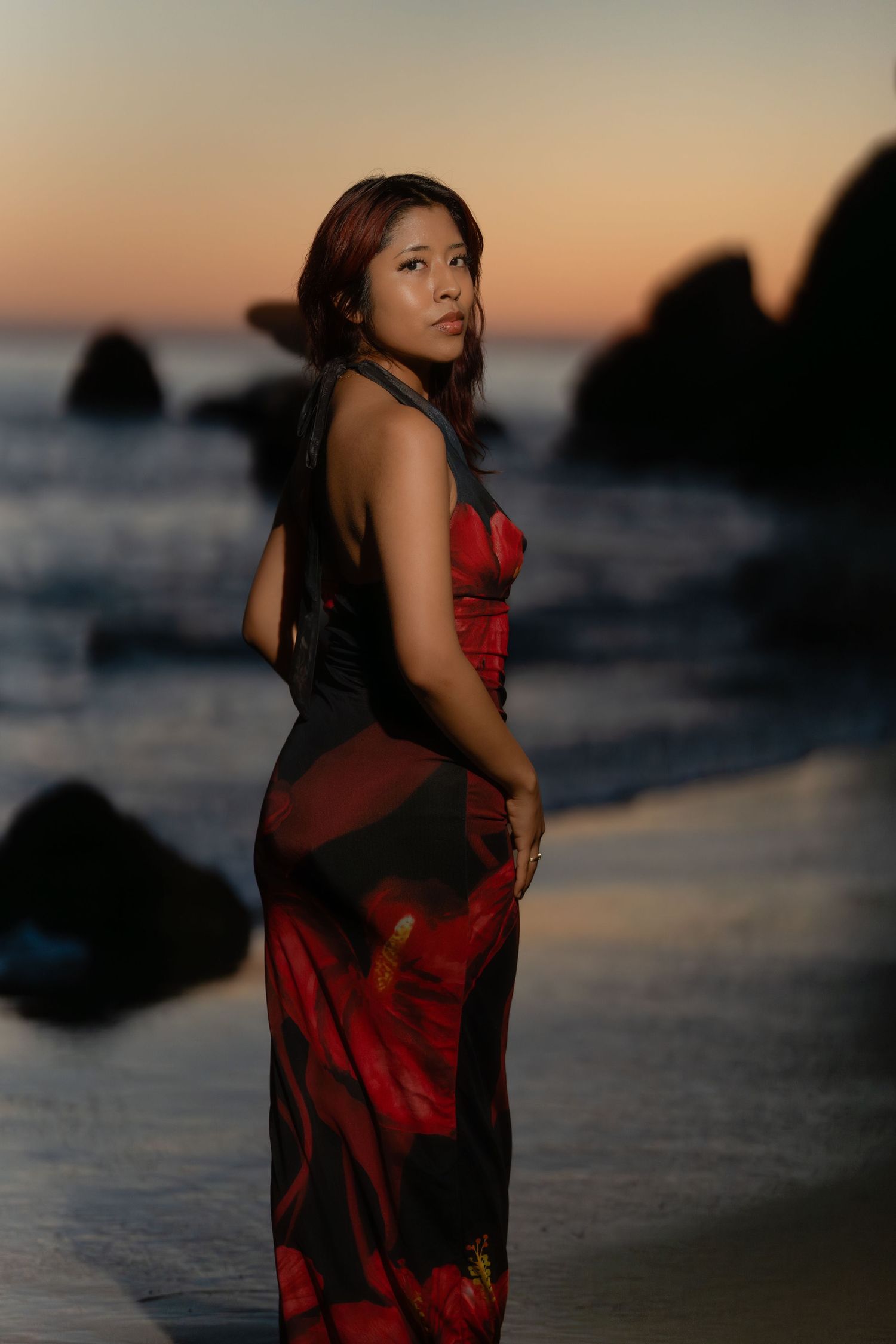 Woman in red floral halter dress posing on beach at sunset with ocean rocks in background.