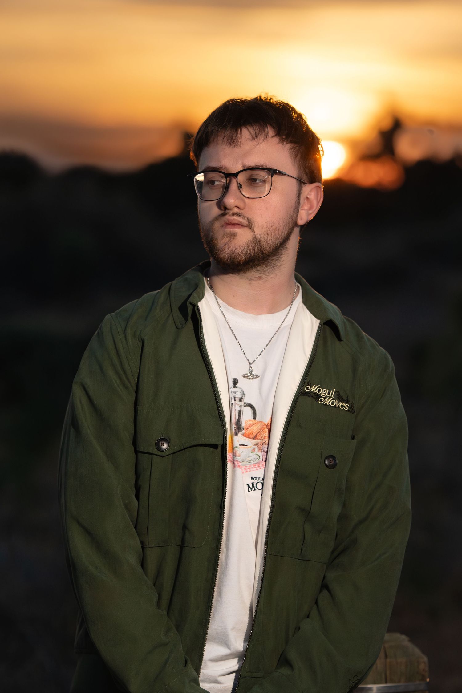 Young man with glasses and green jacket poses outdoors at golden sunset, wearing a white graphic tee and necklace.