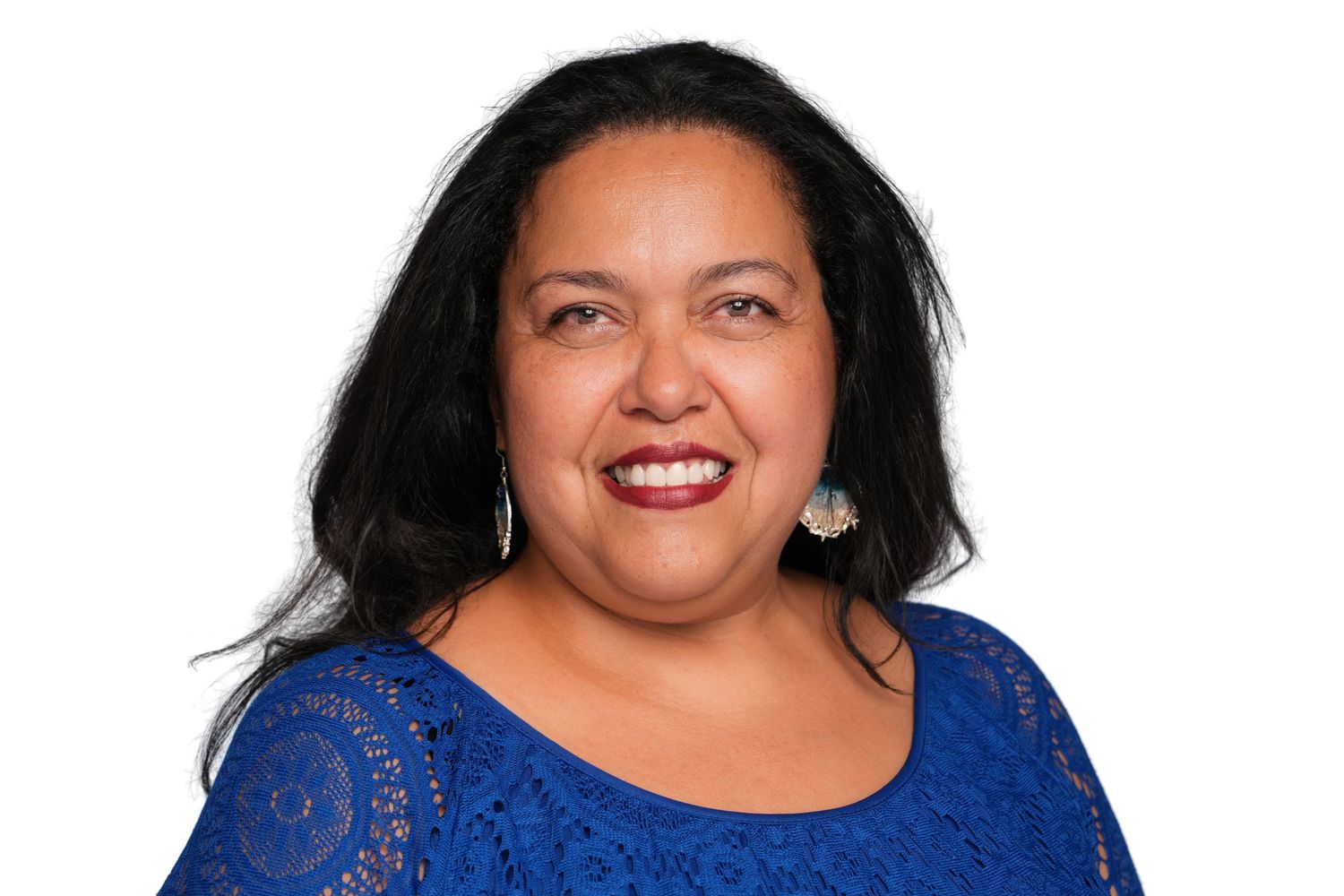 Smiling woman with dark hair wearing a blue lace top and turquoise earrings against a white background.