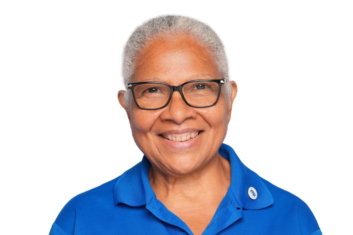 Smiling older woman with short gray hair wearing glasses and a blue polo shirt on a white background.