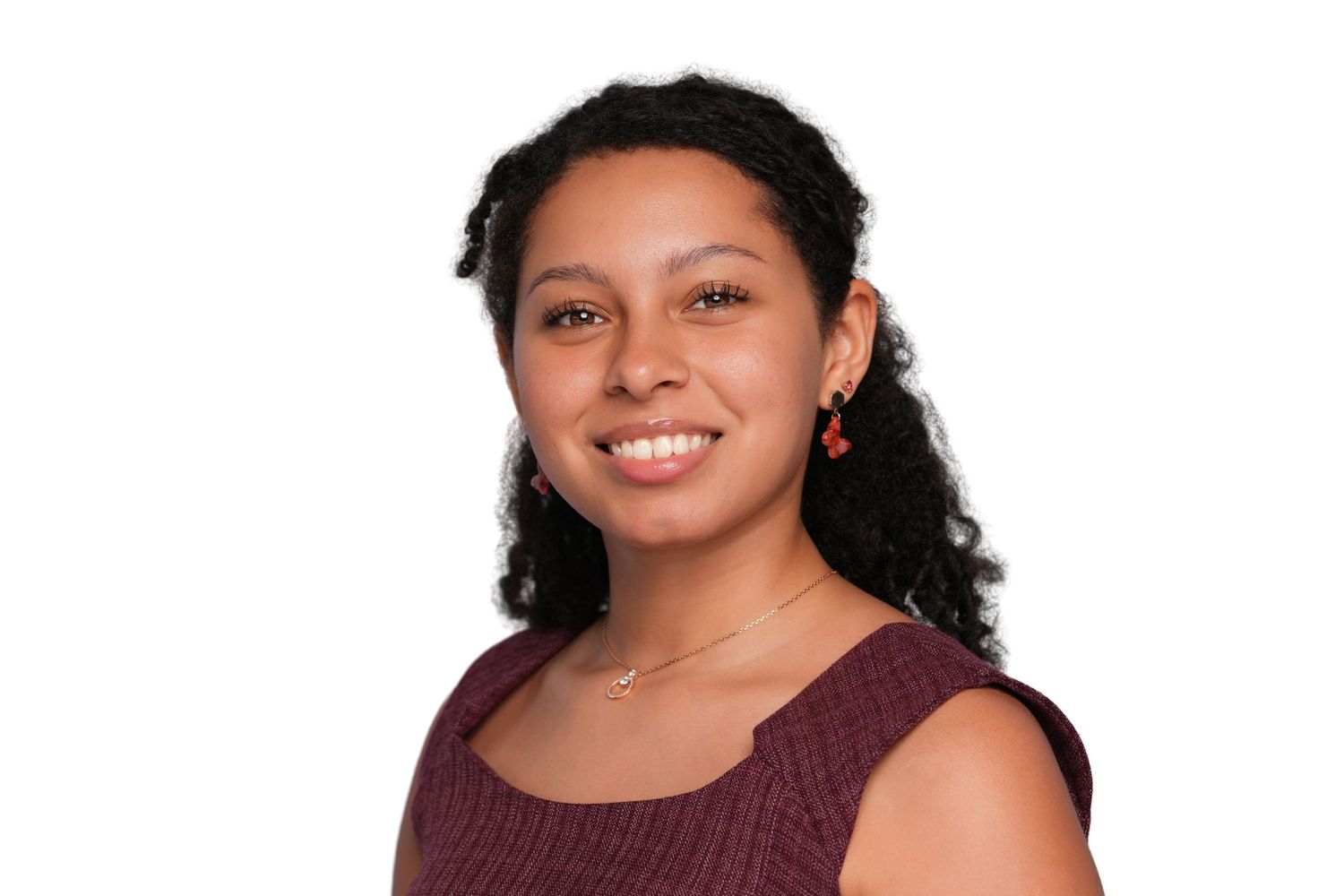 Smiling young woman with curly dark hair wearing a burgundy sleeveless top and red earrings on white background.
