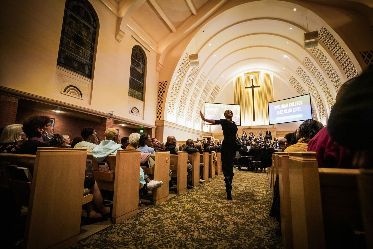 Church choir conductor leads congregation in worship inside ornate chapel with wooden pews and large cross.