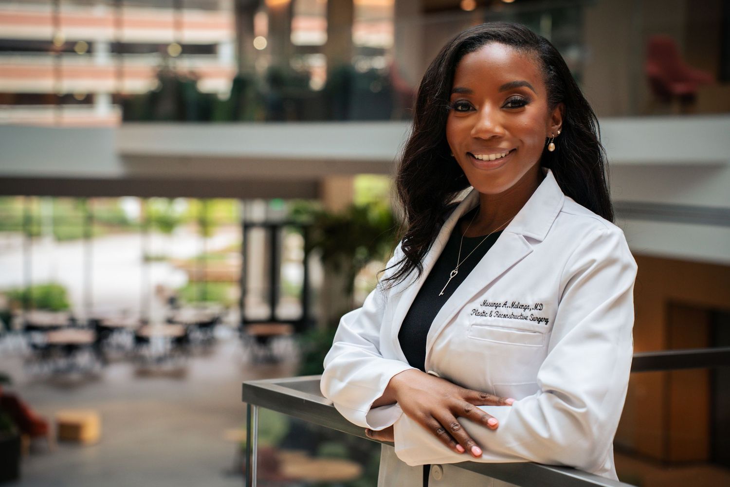 Smiling female doctor in white lab coat poses in bright modern hospital atrium setting.