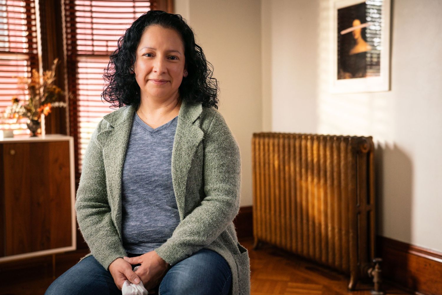 Woman with curly hair sits calmly in a cozy room with a vintage radiator and soft natural light.
