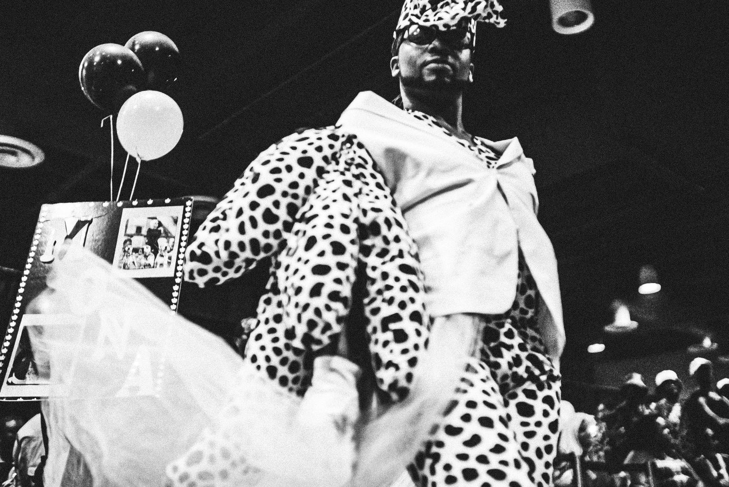 Black and white photo of model in dalmatian print coat and hat posing dramatically at fashion show.