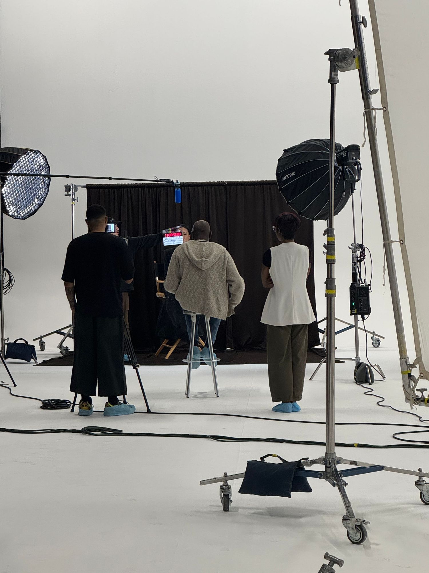 Behind-the-scenes photography studio shoot with crew members, professional lighting equipment, and softboxes on white cyclorama backdrop.