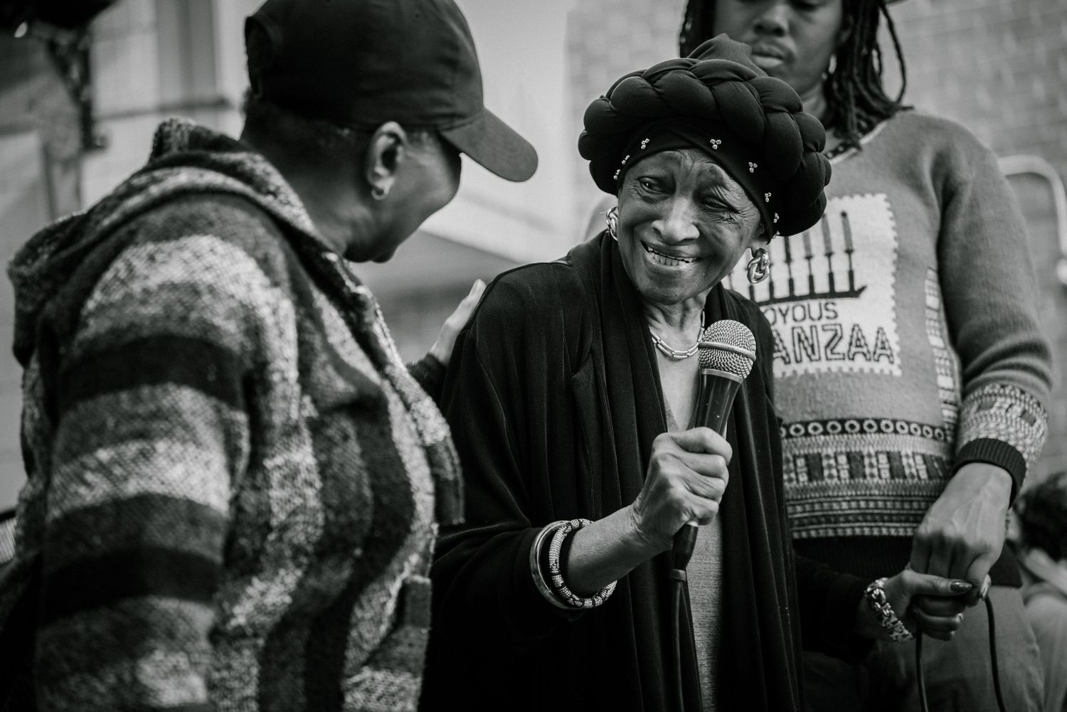 Elderly woman speaks into microphone at outdoor Kwanzaa community event in black and white photo.