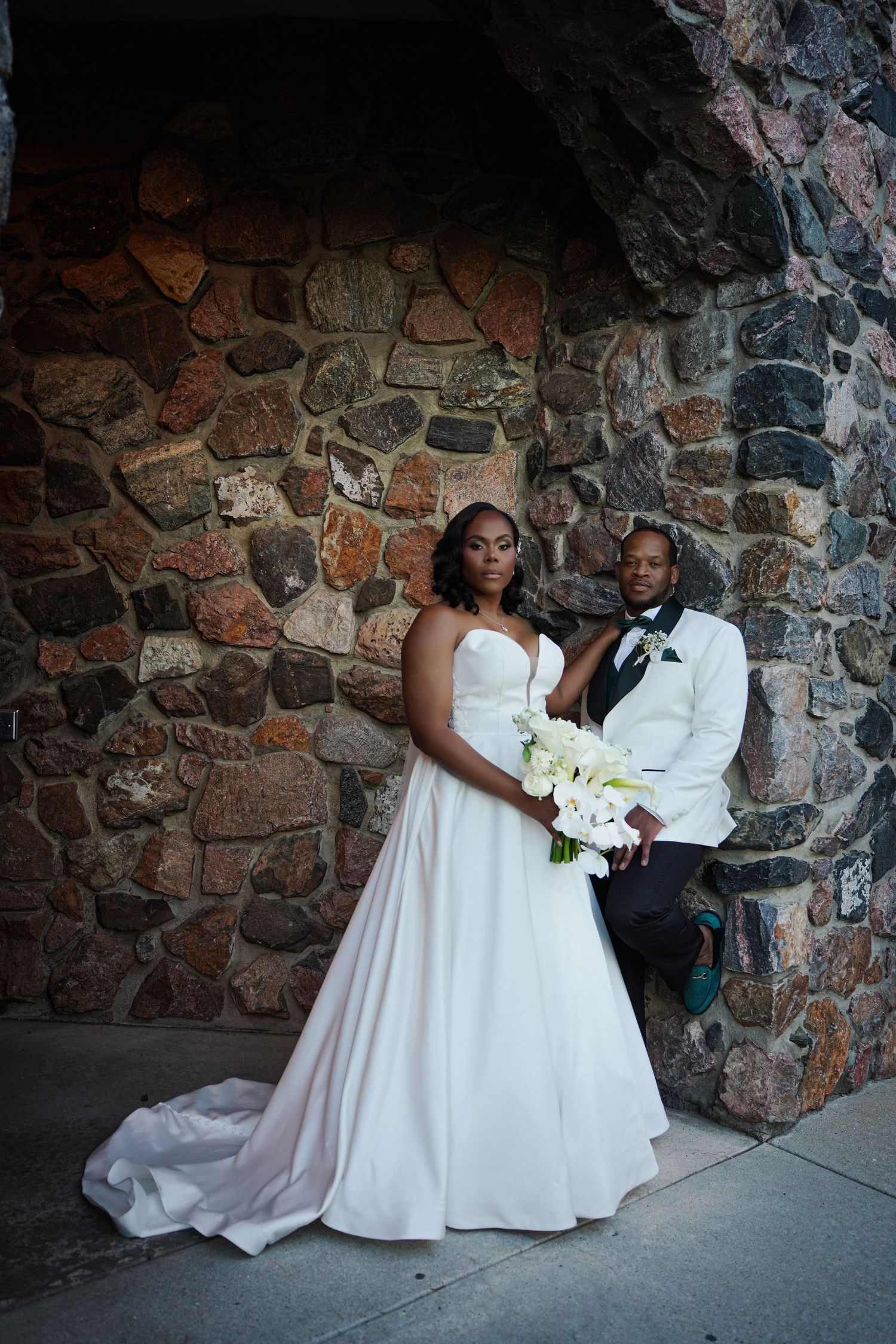 A bride in a white strapless gown holds calla lilies beside a groom in a white tuxedo against a stone wall.