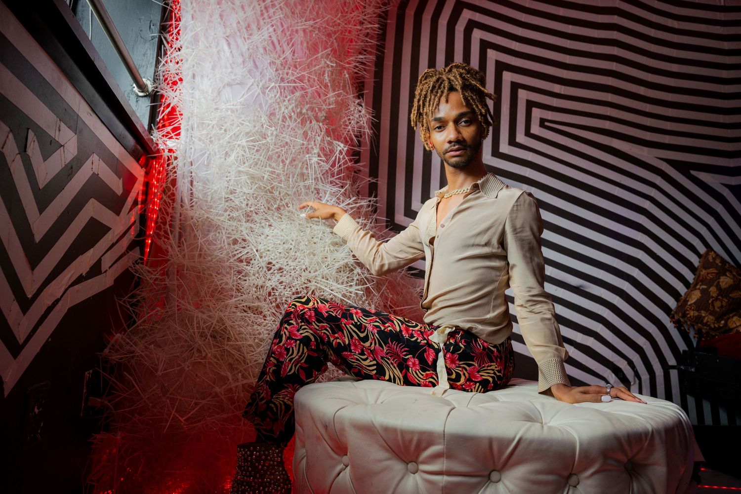 Young man with braids in a beige shirt and patterned pants posing on a white ottoman in a vibrant room.