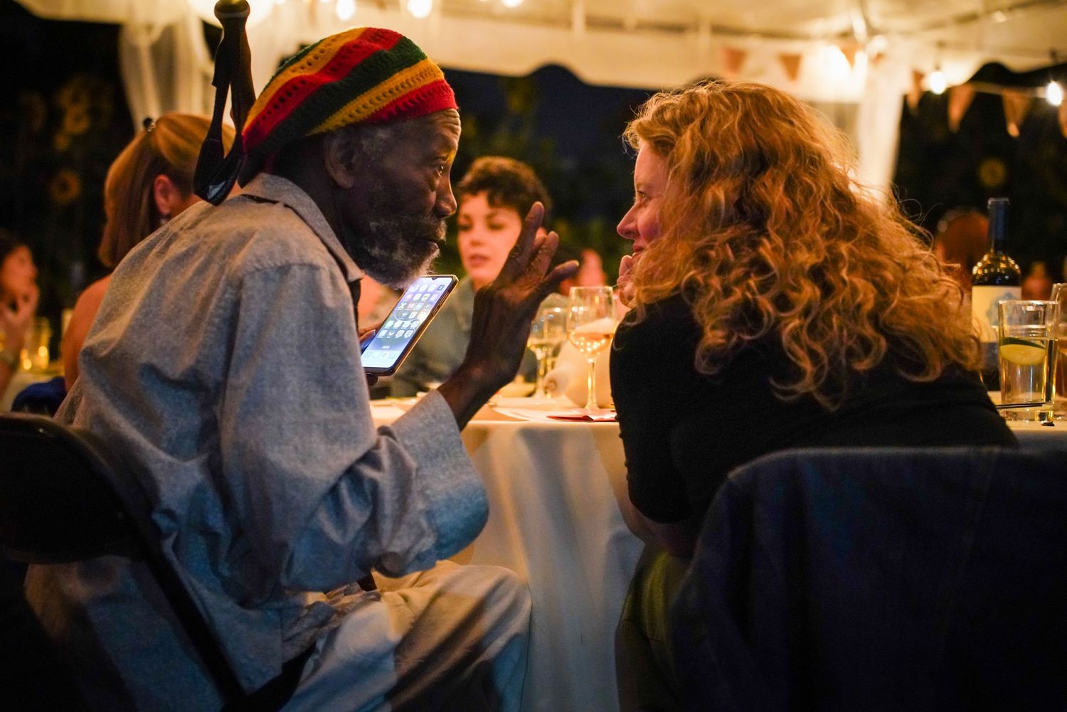 Man in rastafarian hat showing phone to curly-haired woman at outdoor dinner party.
