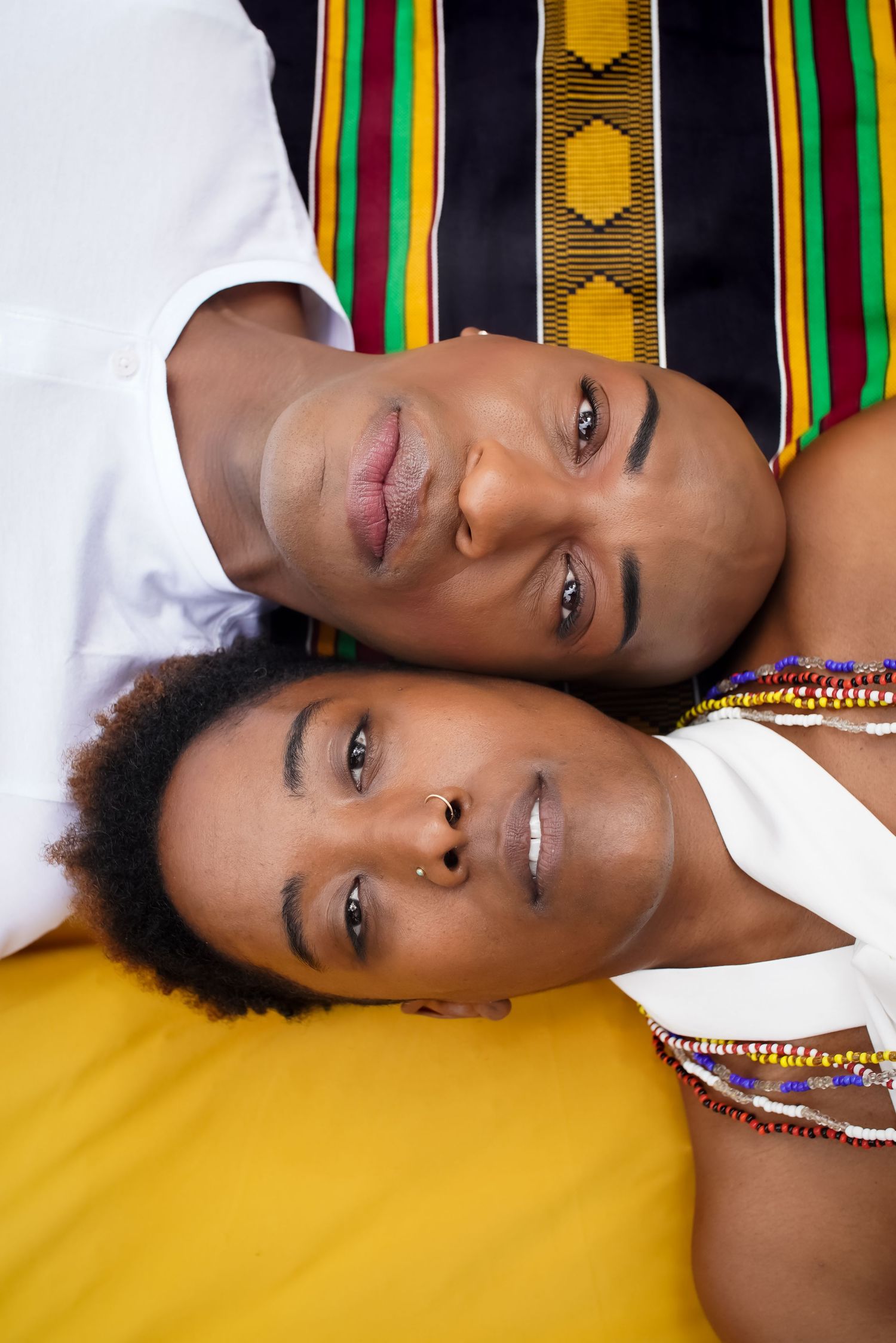 Two Black individuals wearing traditional African Kente cloth and beaded jewelry lie side by side looking up at the camera.