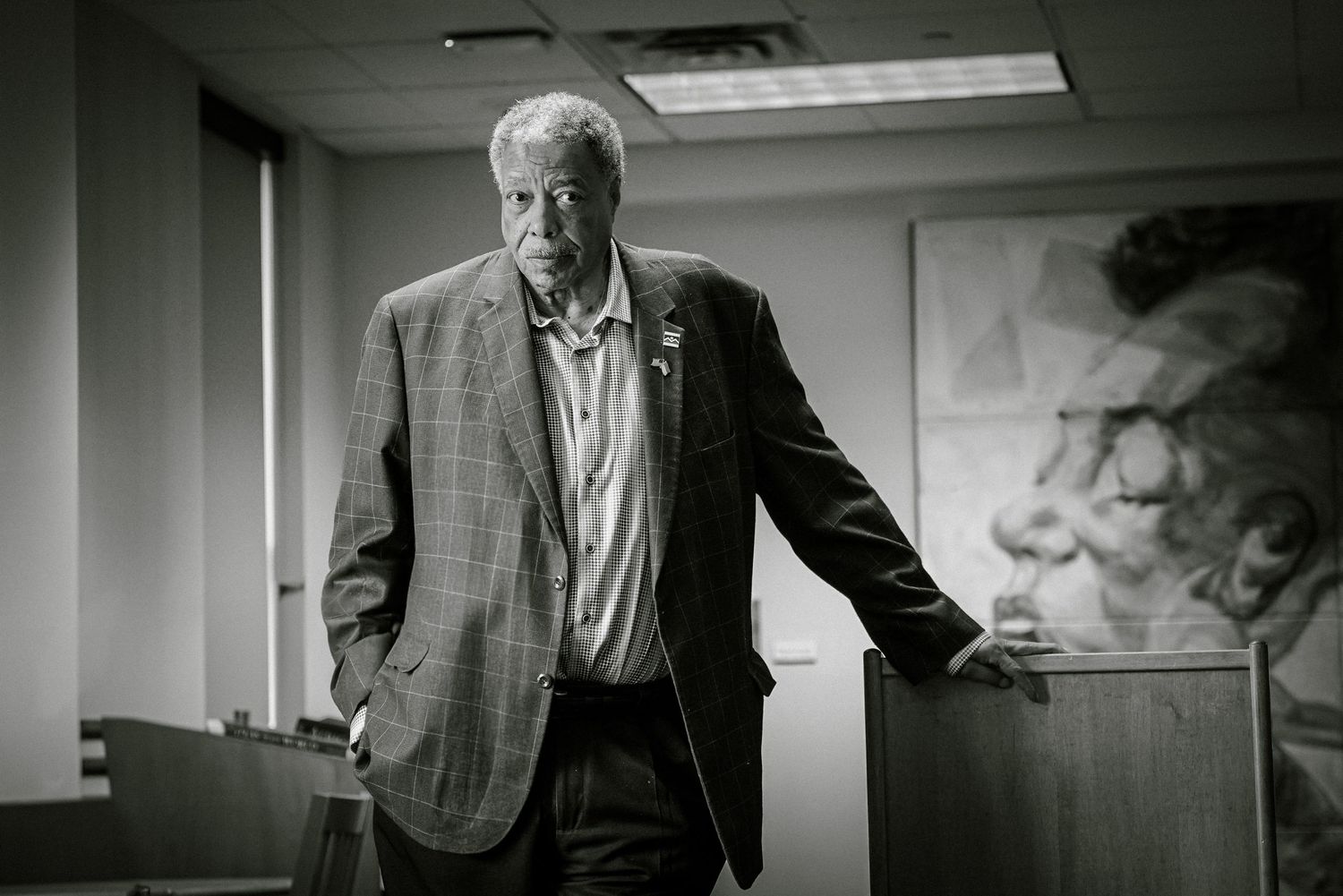 Black and white portrait of distinguished older man in blazer leaning on podium beside large artwork.