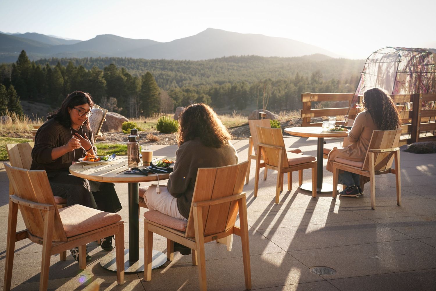 Three women dining outdoors on mountain terrace with scenic forest and sunset views in background.