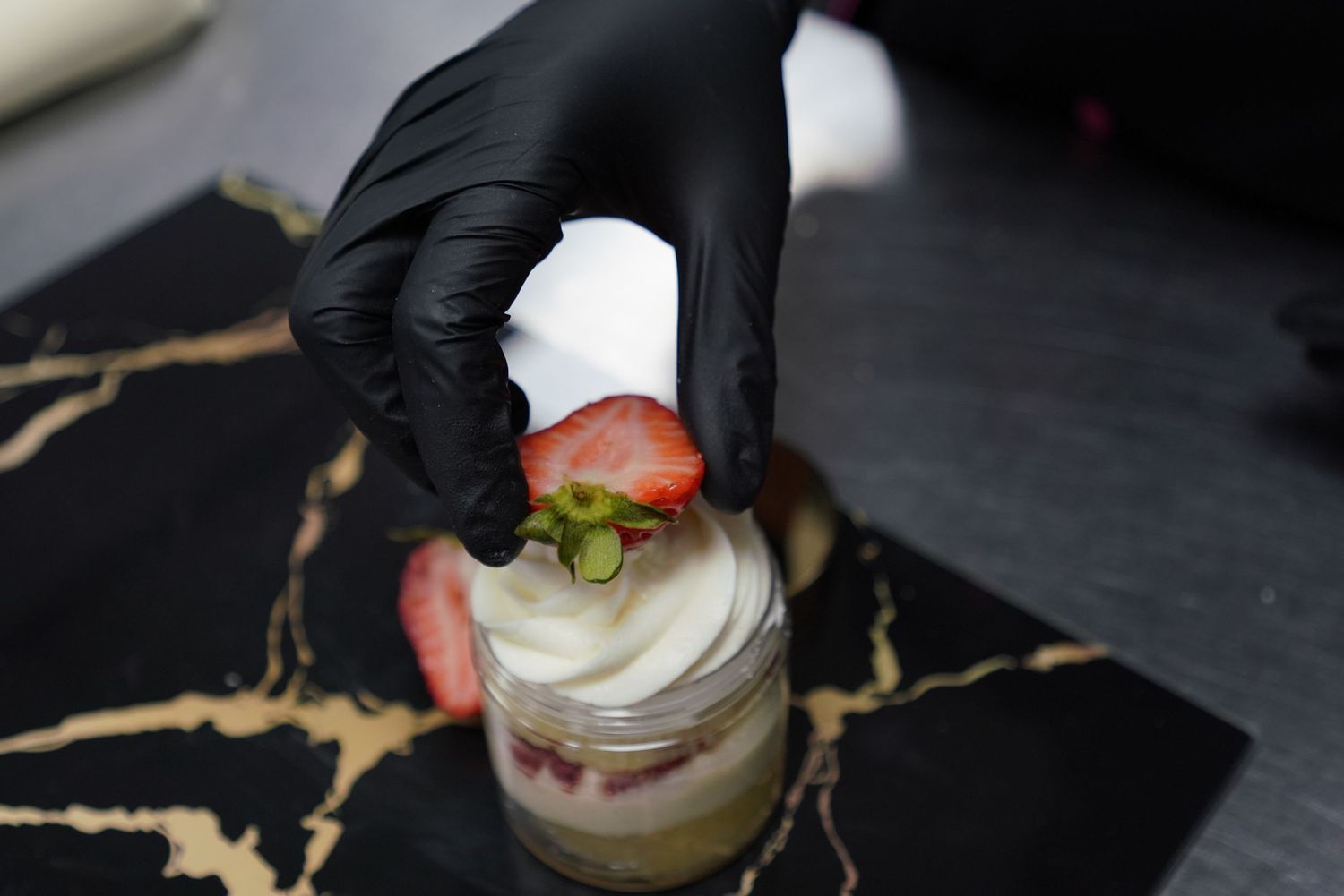 Gloved hand topping a creamy dessert cup with a fresh strawberry slice on a black and gold marble serving board.