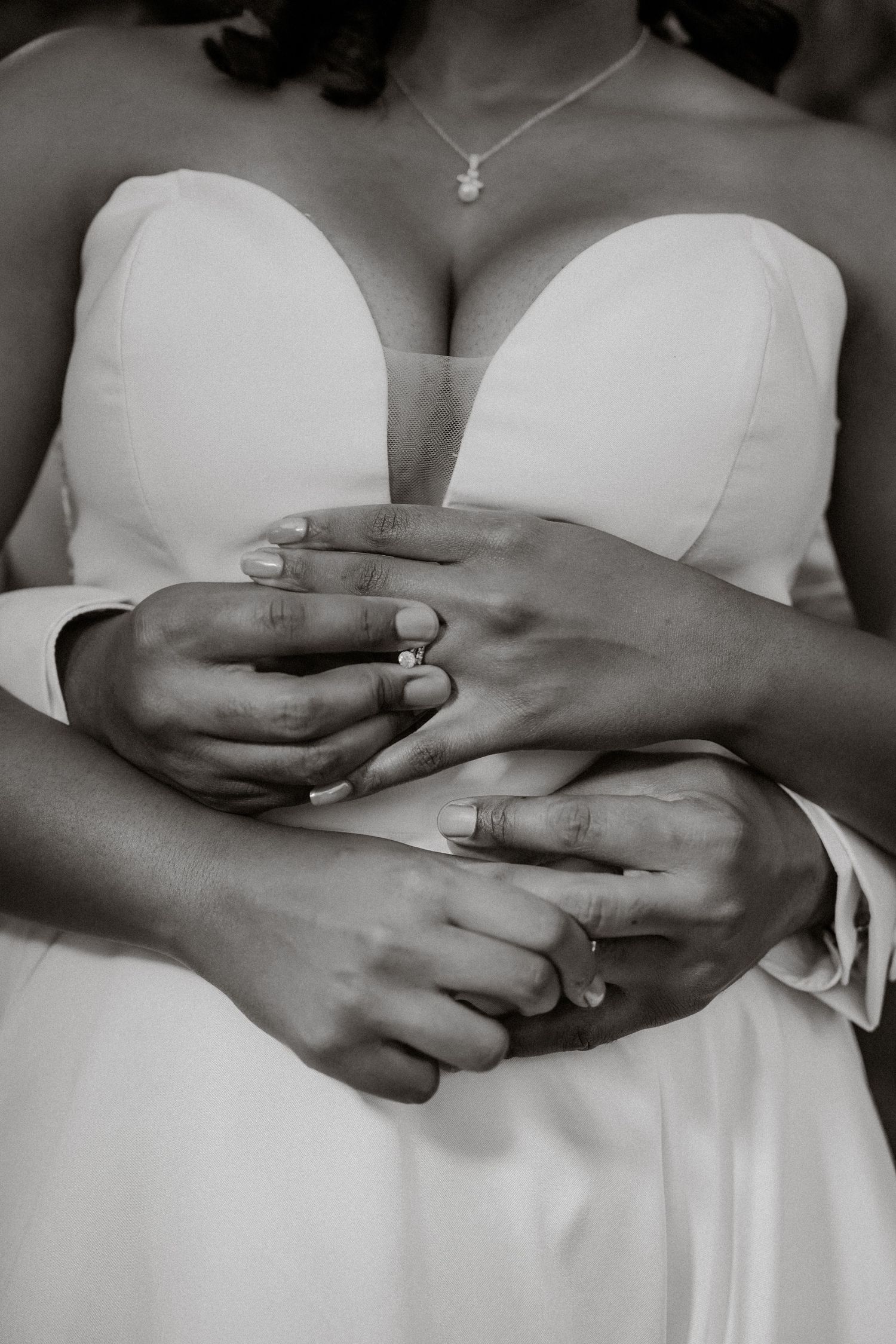 Black and white photo of newlyweds hands intertwined showing wedding rings, bride in strapless gown.