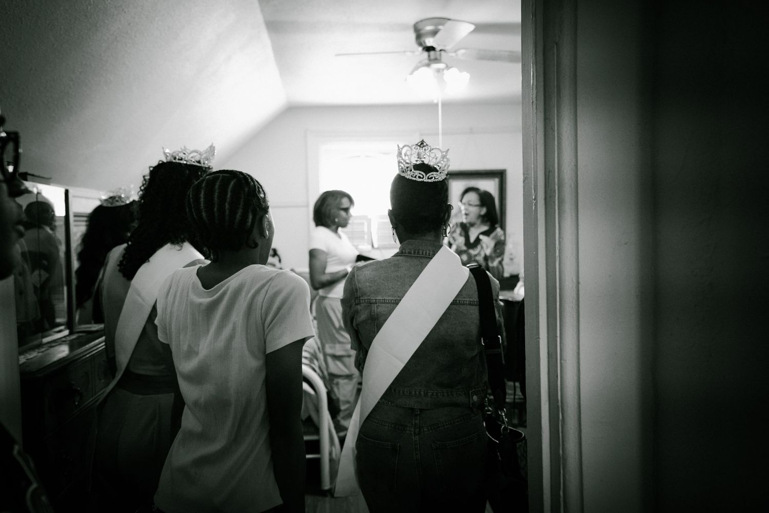 Black and white photo of women wearing crowns and sashes in a backstage dressing room.