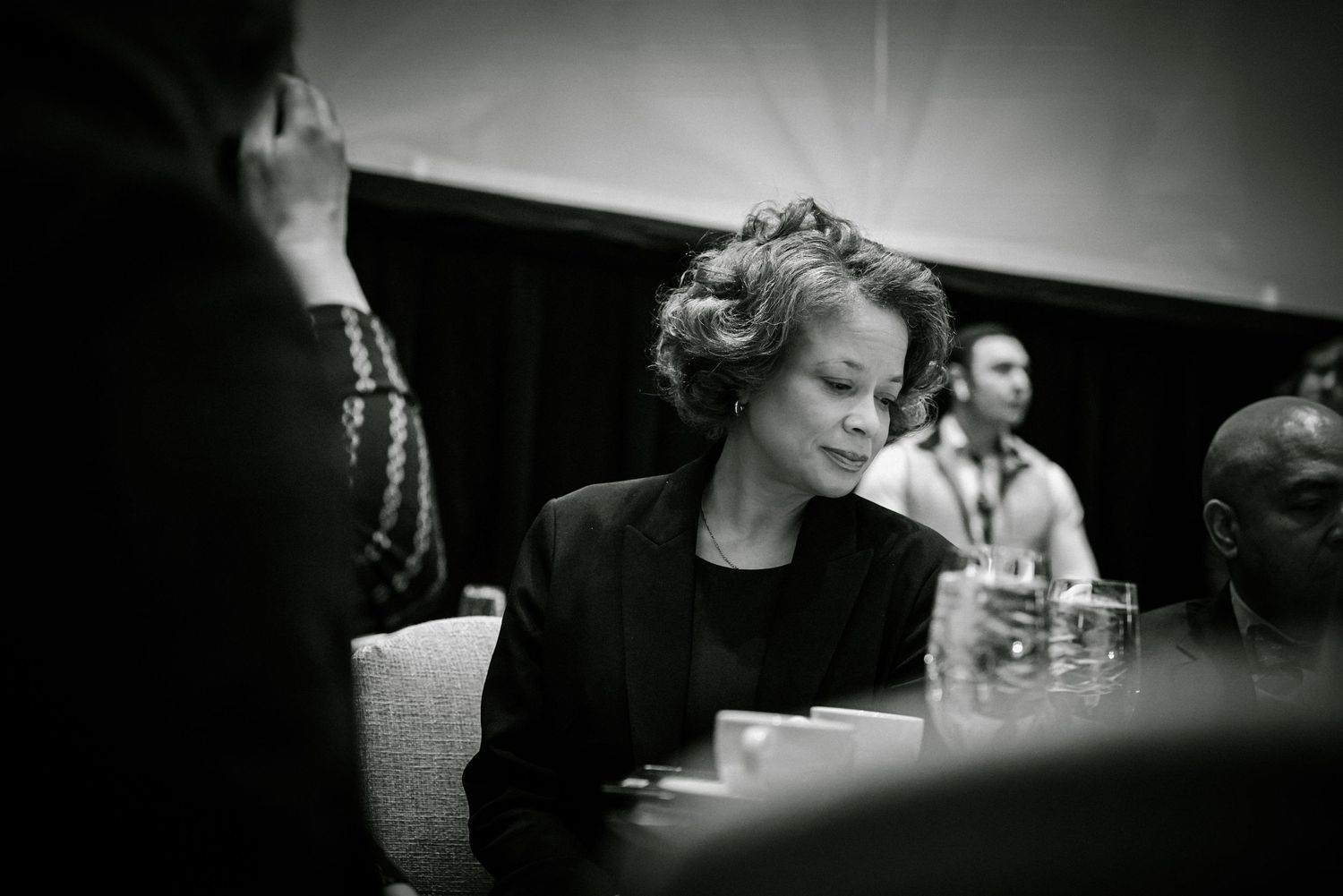 Black and white candid photo of woman with short hair seated at conference table during formal meeting.