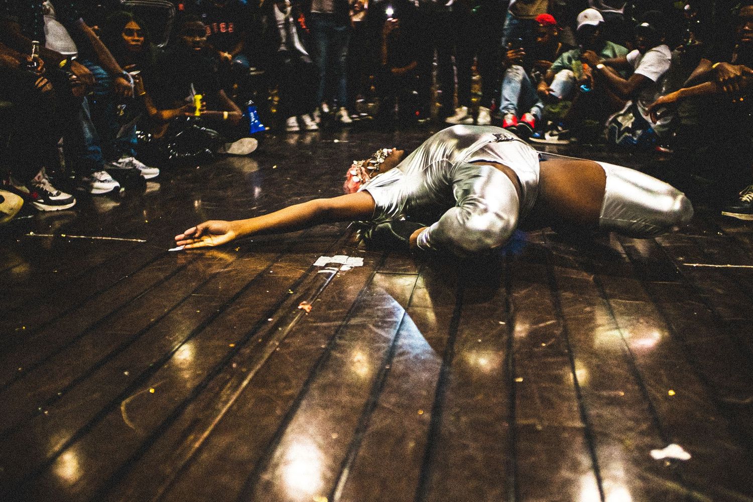 A performer in silver outfit lies dramatically outstretched on a wooden dance floor at a nightclub.