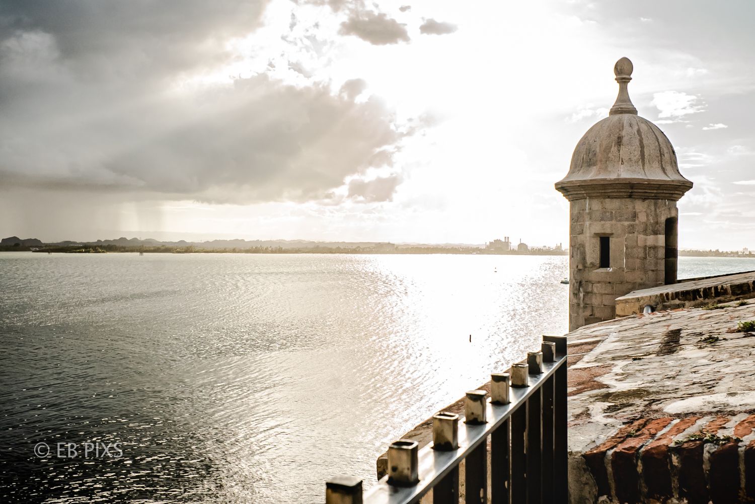 Historic stone watchtower of El Morro fort overlooks calm harbor waters under dramatic cloudy sky in San Juan, Puerto Rico.
