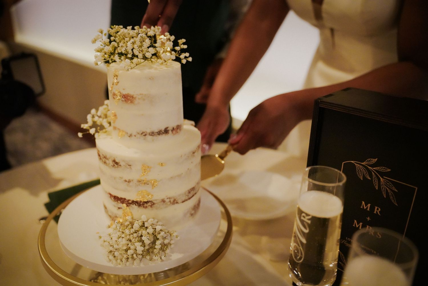 Couple cutting a three-tier semi-naked wedding cake decorated with baby's breath flowers on a gold stand.