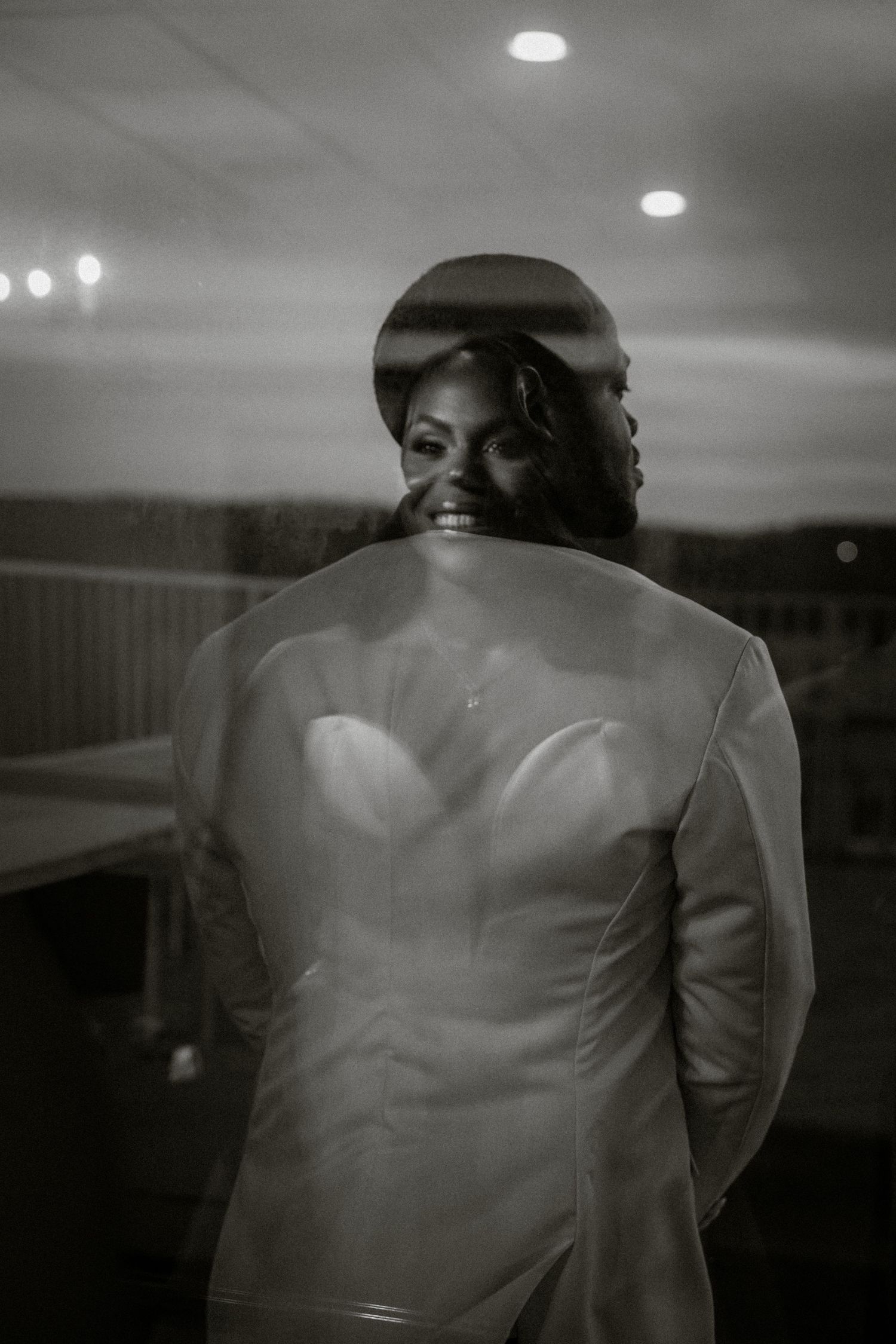 Black and white double exposure wedding photo of a smiling bride in a strapless gown with her groom reflected in glass.