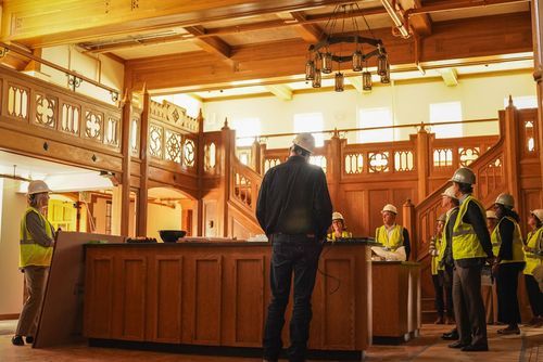 Construction workers in hard hats inspect ornate wooden interior of historic building renovation.