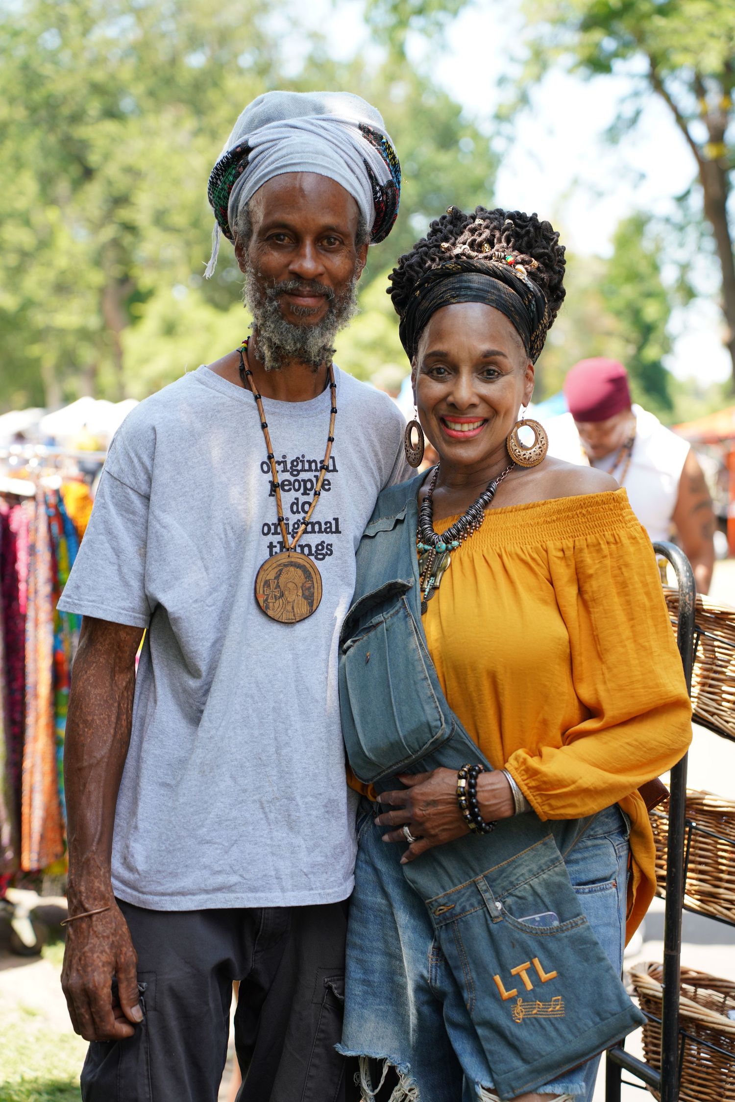 Couple poses together outdoors at market, man in gray tee with head wrap, woman in yellow off-shoulder top.