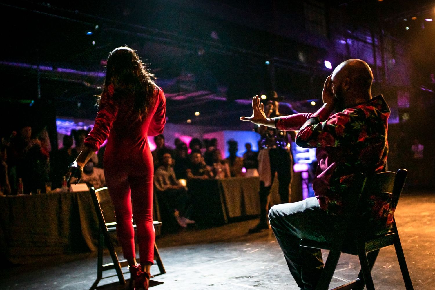Musician plays violin on stage while performer in red suit stands under pink spotlight at live event.
