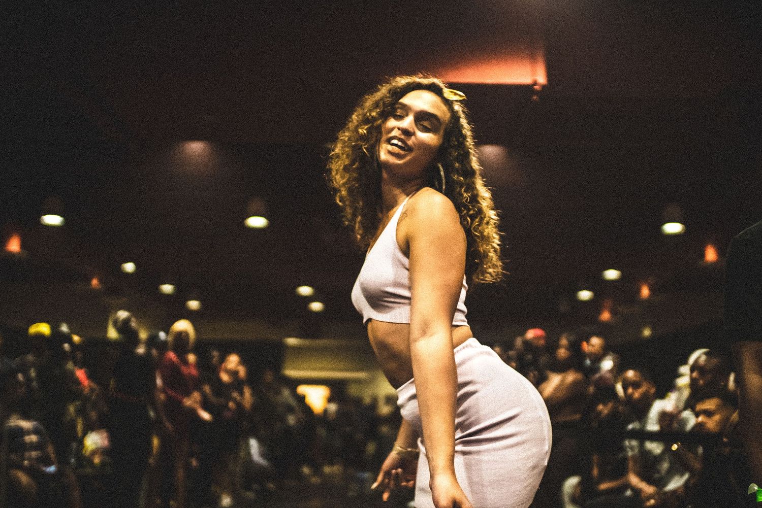 Young woman with curly hair in gray two-piece outfit poses confidently at indoor event.