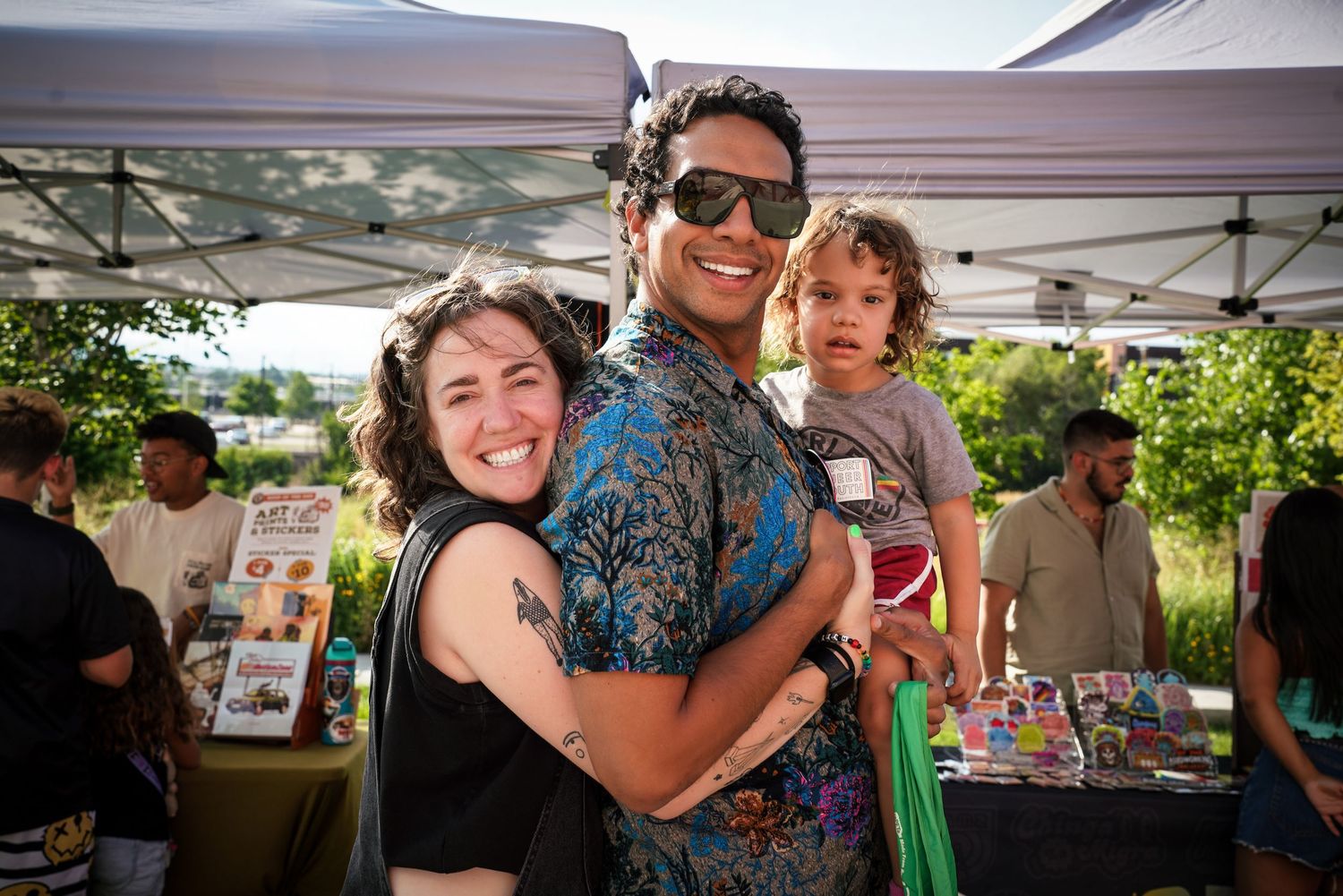 Two adults and a child share a warm hug at an outdoor Pride community festival.
