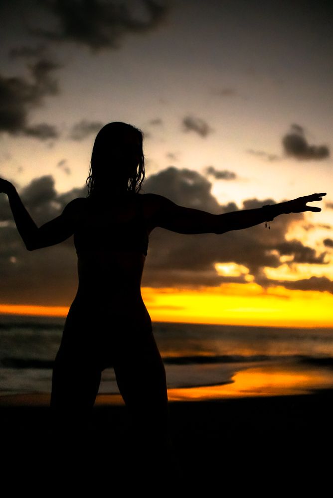Close-up silhouette of a woman with arms spread wide against a vivid orange and purple sunset sky.