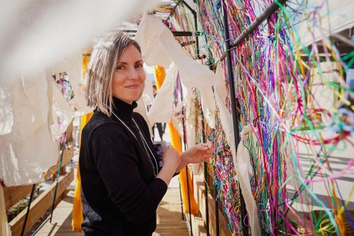 Female artist examines colorful hanging textile installation in a bright creative studio space.