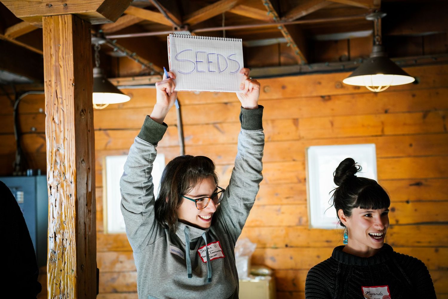 Young girl enthusiastically holds up a handwritten SEEDS sign overhead while smiling in a rustic wooden cabin setting.