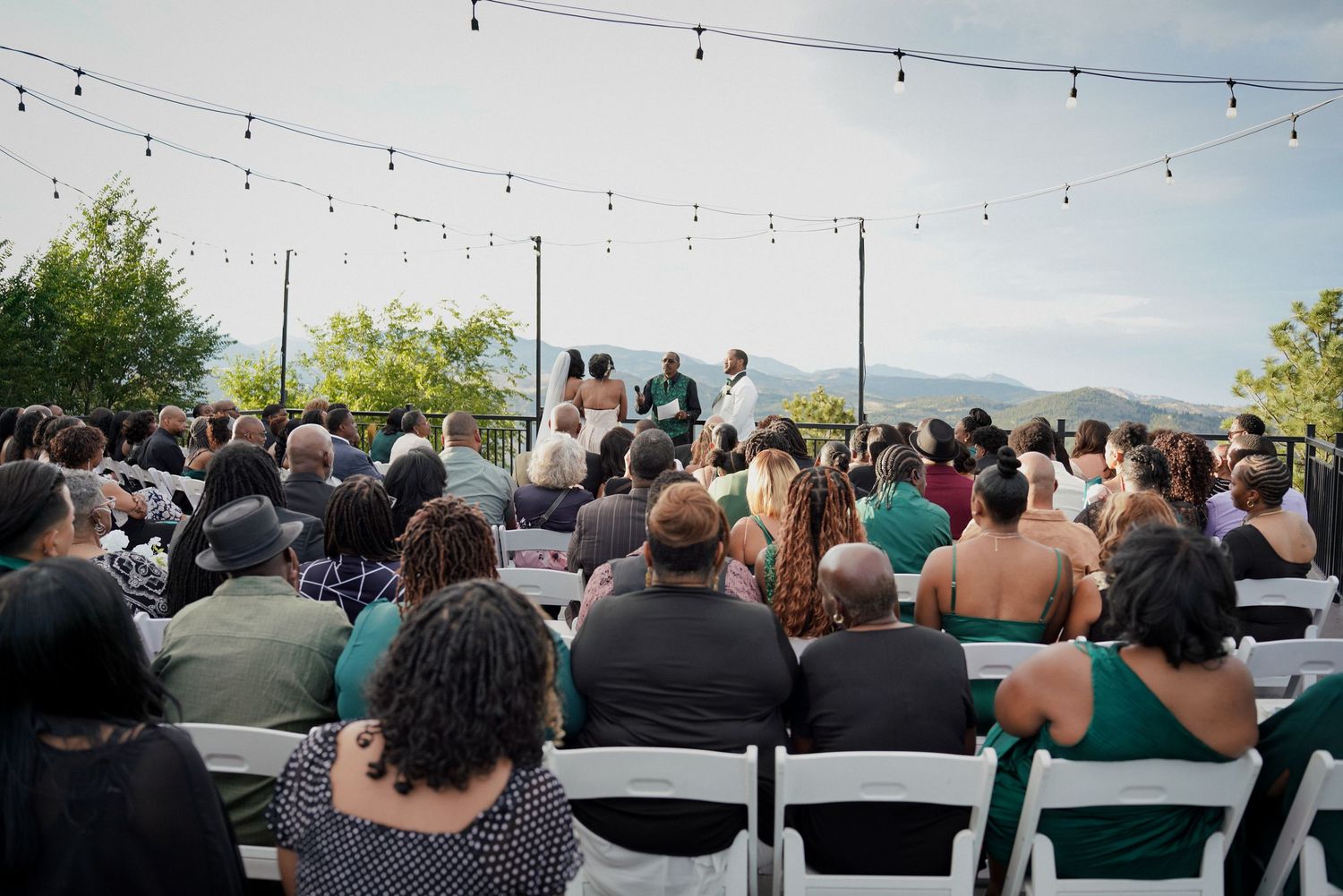 Outdoor wedding ceremony with guests seated in white chairs under string lights with mountain views.