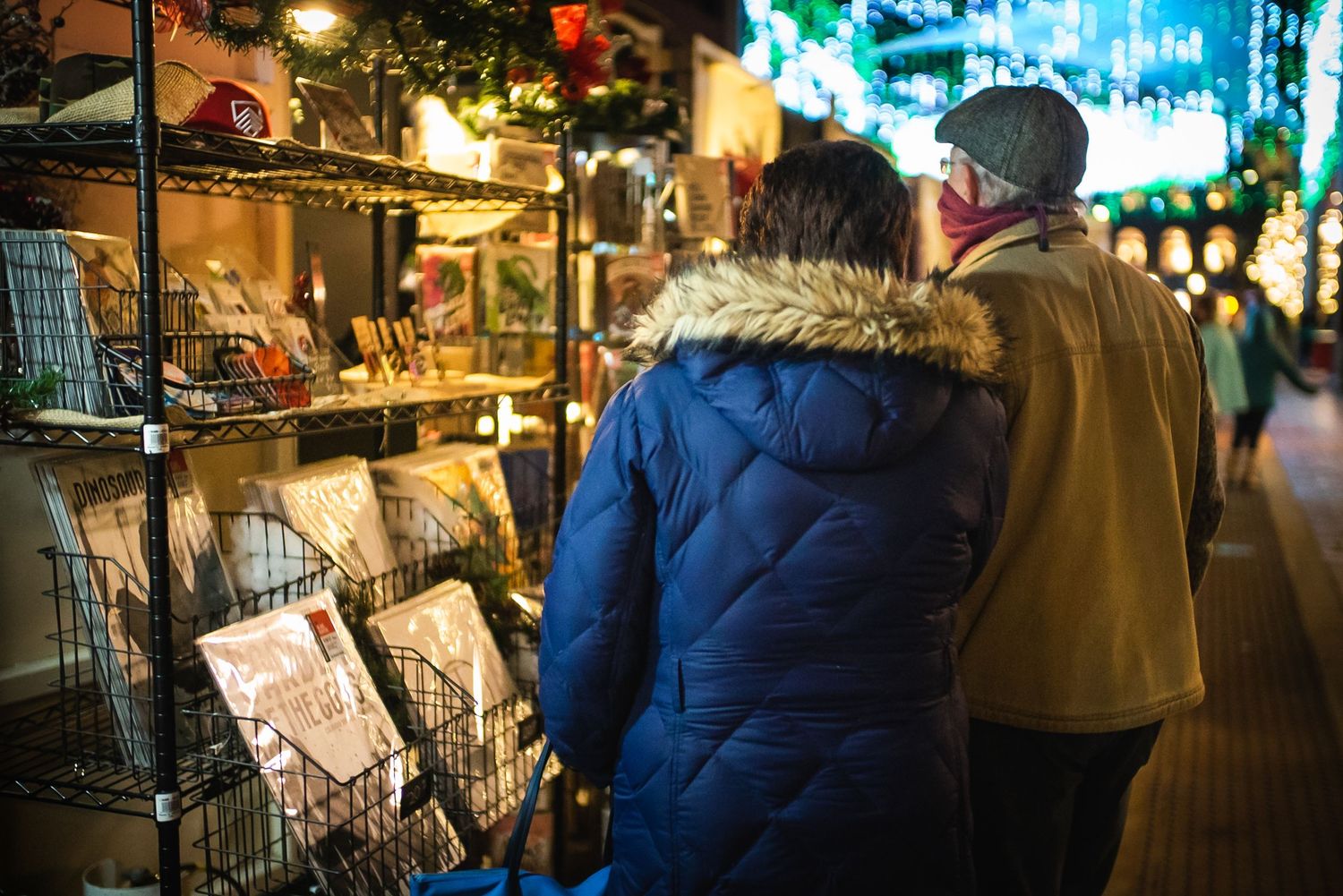 A couple in winter coats browsing an outdoor market stall decorated with Christmas lights.