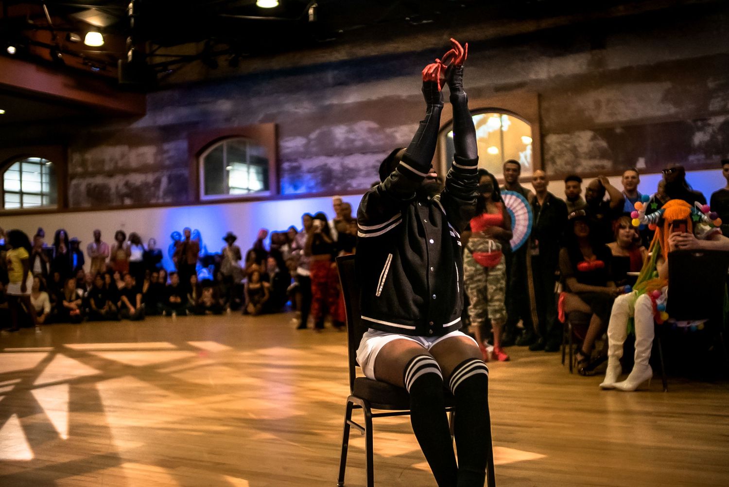 A performer in thigh-high socks raises legs while seated on a chair at a dance event.