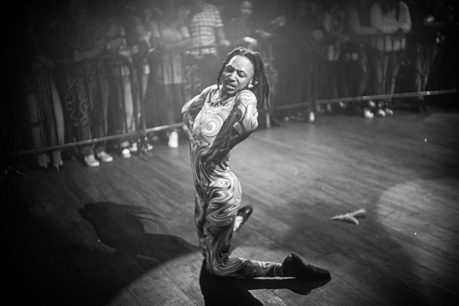 Black and white photo of dreadlocked dancer on knees performing expressively on stage spotlight.