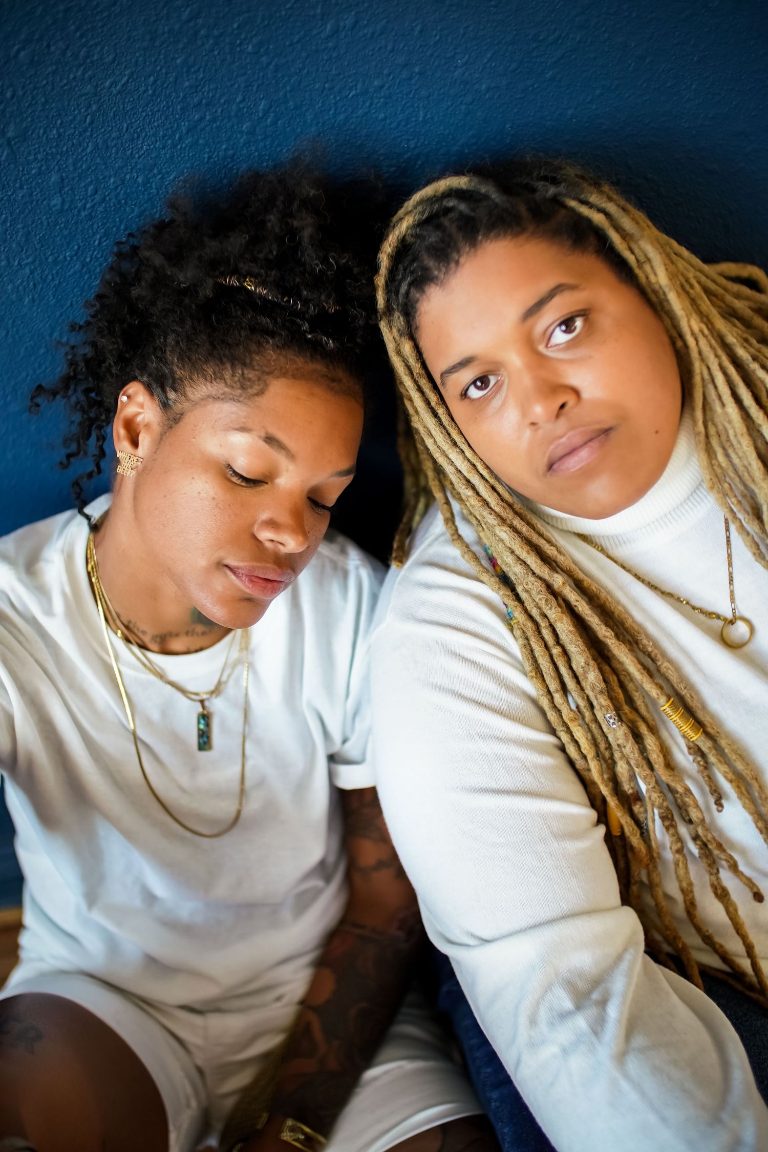 Two people in white outfits sit against a blue wall, one with curly hair and one with blonde dreadlocks, wearing gold necklaces.