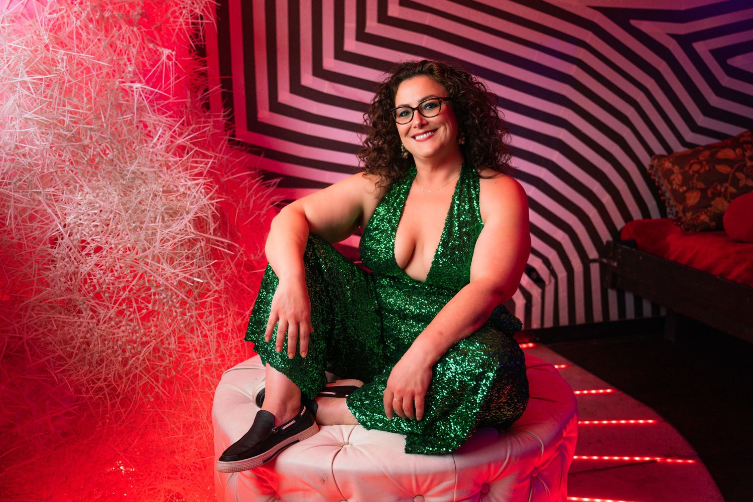 A woman in a green sequined dress sits smiling on a white ottoman surrounded by red lights and geometric patterned walls.