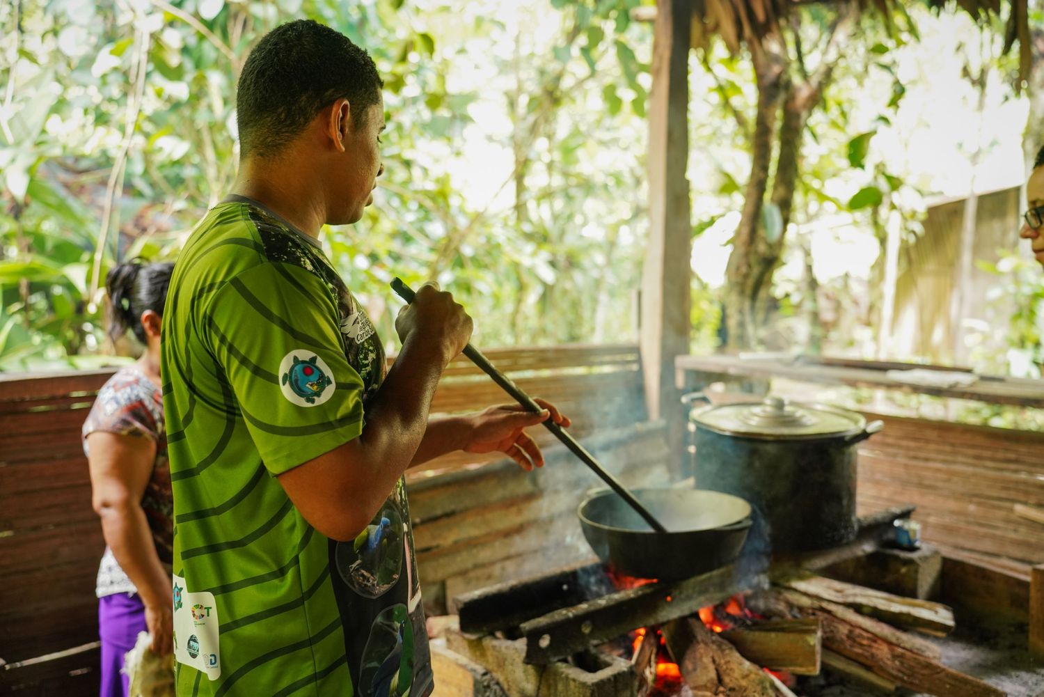 Man cooking over open fire in rustic outdoor kitchen surrounded by tropical jungle foliage.