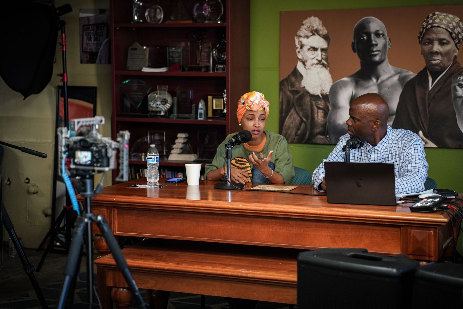 Two people conduct recorded podcast interview at wooden desk with camera and microphone setup indoors.