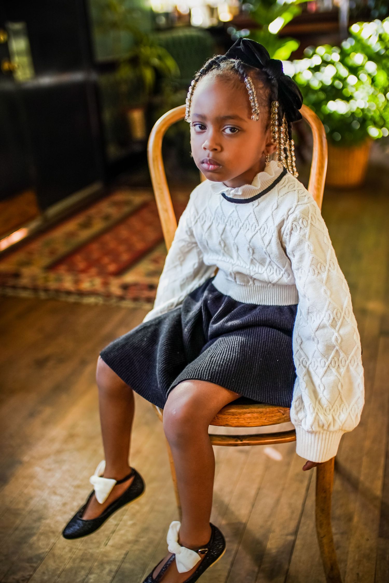 Young girl in white sweater and gray skirt seated on wooden chair.