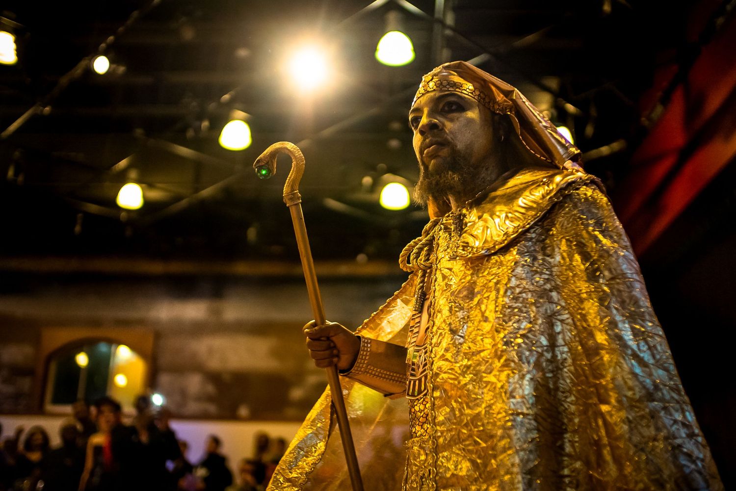 A performer in an elaborate gold cape and crown holds a staff under dramatic stage lighting.